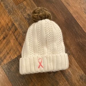 Cream colored beanie with pink ribbon on it and poof ball on the top